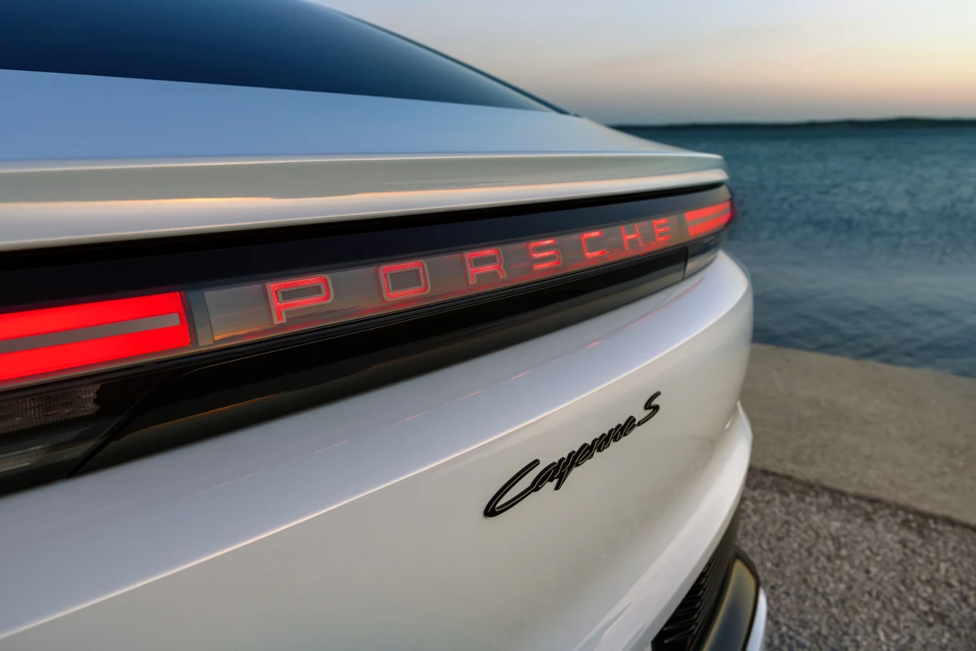 tylny fragment białego Porsche Carrera S z podświetlanym czerwonym napisem marki na listwie świetlnej, ustawiony nad wodą o zmierzchu