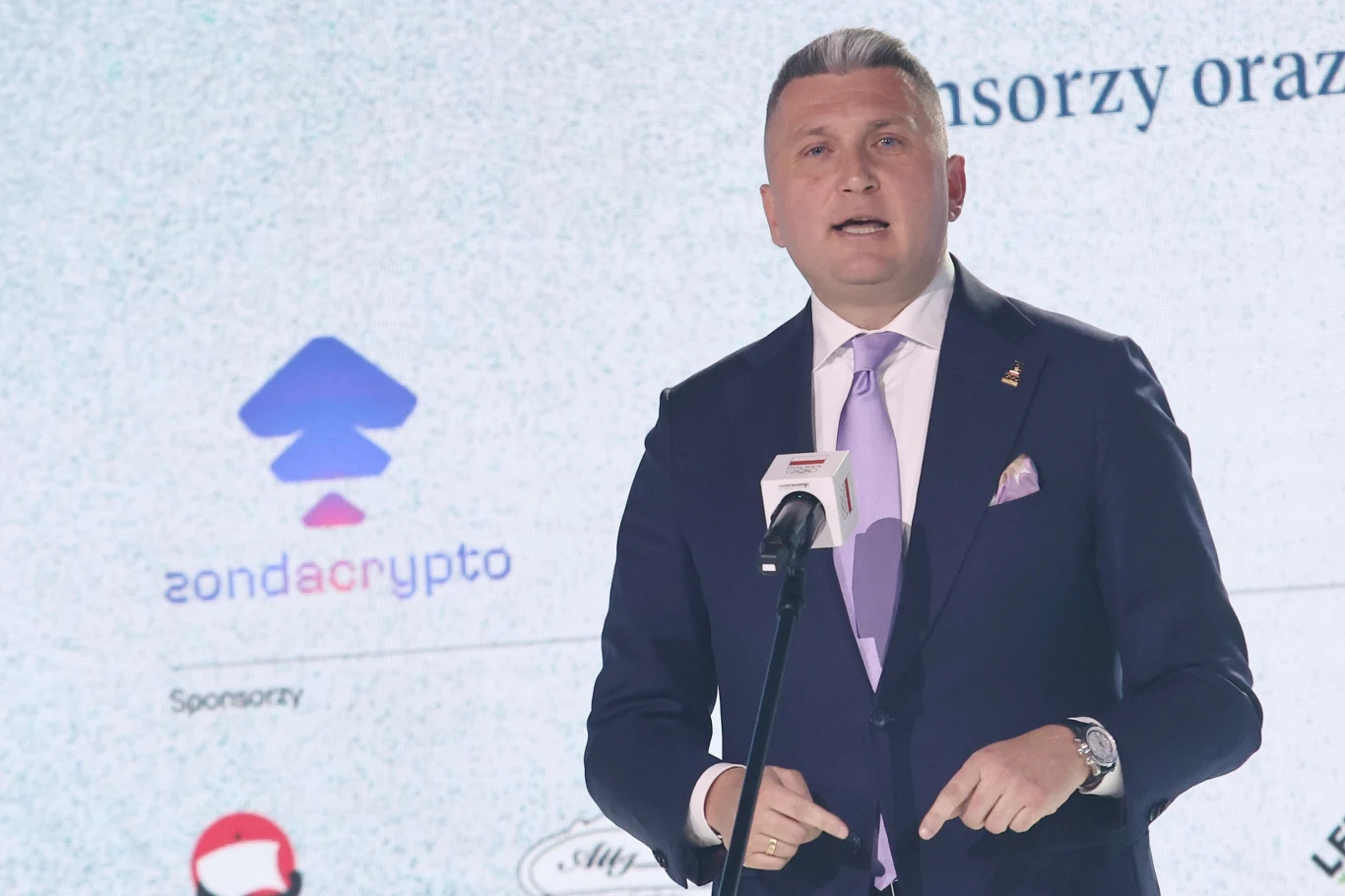 Radosław Piesiewicz Radosław Piesiewicz w garniturze i fioletowym krawacie przy mikrofonie na tle logotypu zondacrypto.