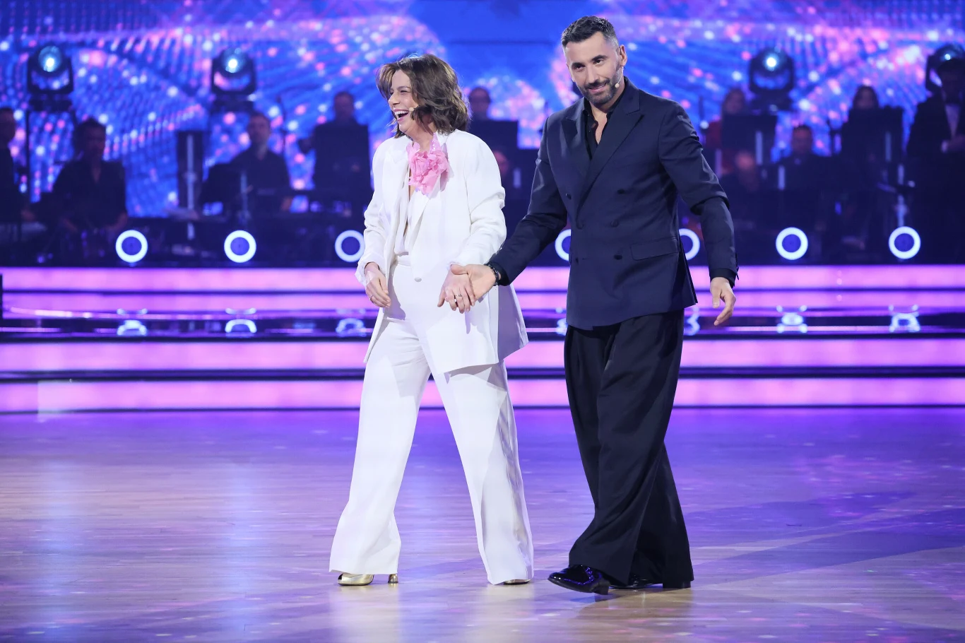 Para tancerzy trzyma się za ręce na parkiecie, ubrana elegancko; kobieta w białym garniturze z różowymi akcentami, mężczyzna w ciemnym, szerokim garniturze, w tle kolorowe światła i zarys orkiestry.