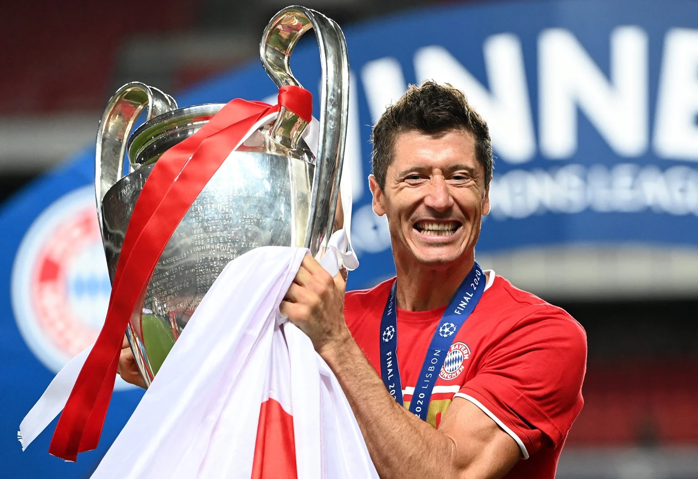 piłkarz w czerwonej koszulce drużyny z medalem na szyi trzyma w rękach dużą srebrną trofeum ozdobione białą i czerwoną wstążką, na jego twarzy widać szeroki uśmiech zdradzający radość ze zwycięstwa
