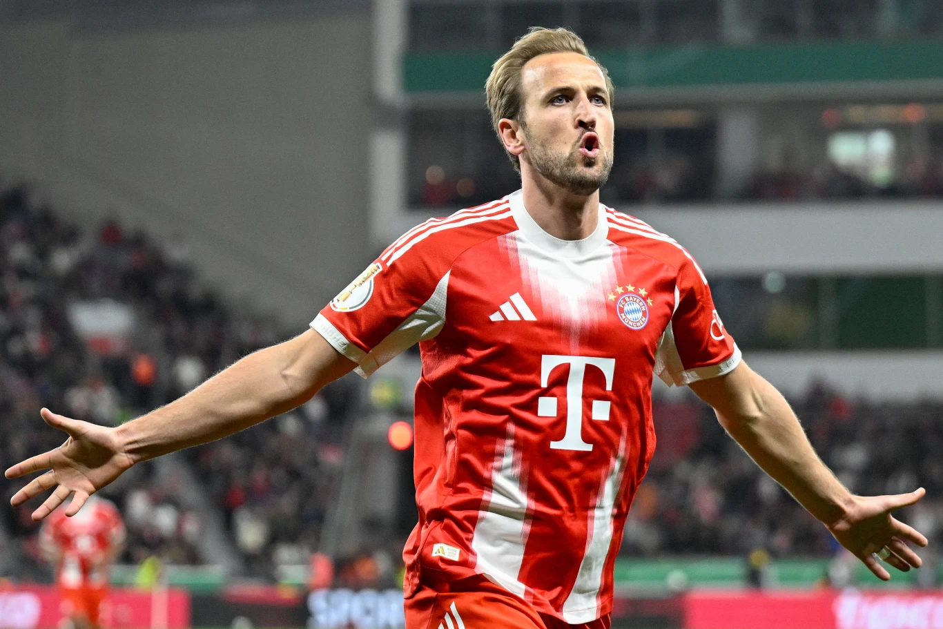 piłkarz w czerwono-białym stroju Bayernu Monachium z rozłożonymi ramionami świętuje zdobycie bramki na tle stadionu wypełnionego kibicami