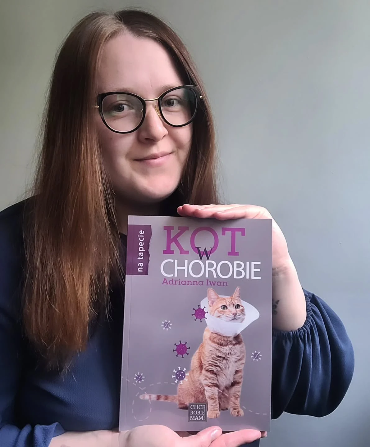 Książka Adrianny Iwan skierowana jest do opiekunów kotów Kobieta w okularach trzyma w rękach książkę zatytułowaną „Kot w chorobie” z ilustracją kota w elżbietańskim kołnierzu na okładce.
