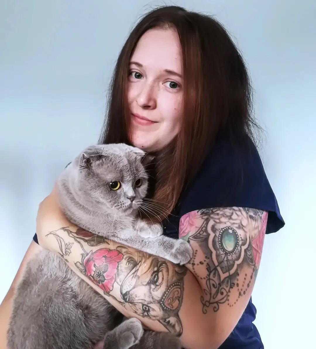 "Koty są mistrzami w maskowaniu bólu" - mówi Adrianna Iwan kobieta z długimi brązowymi włosami w granatowej koszulce trzyma na rękach szarego kota o żółtych oczach; na jej rękach widać liczne, kolorowe tatuaże