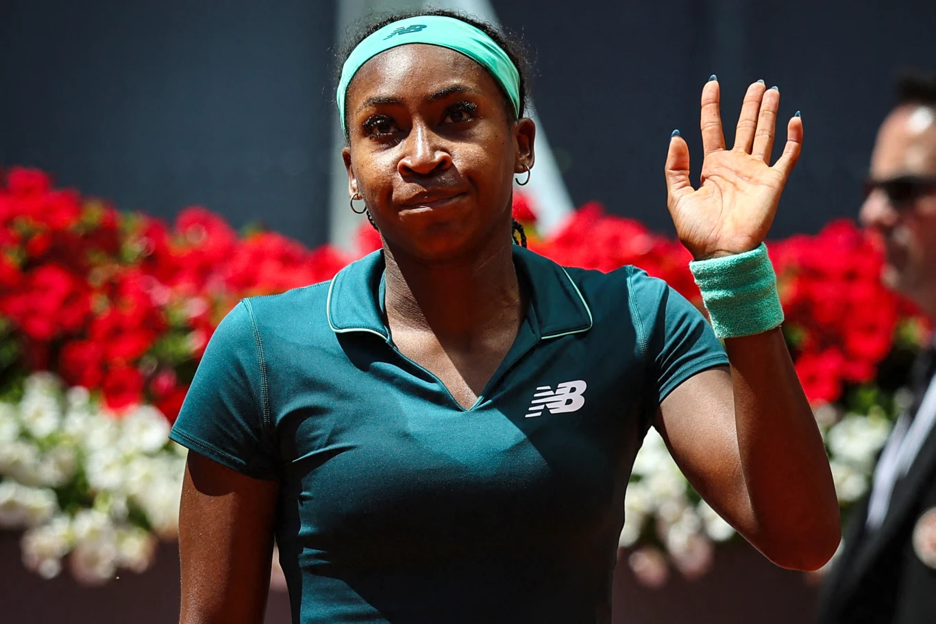 tenisistka w zielonym stroju sportowym oraz opasce na włosach unosi rękę w geście pozdrowienia, w tle czerwone i białe kwiaty