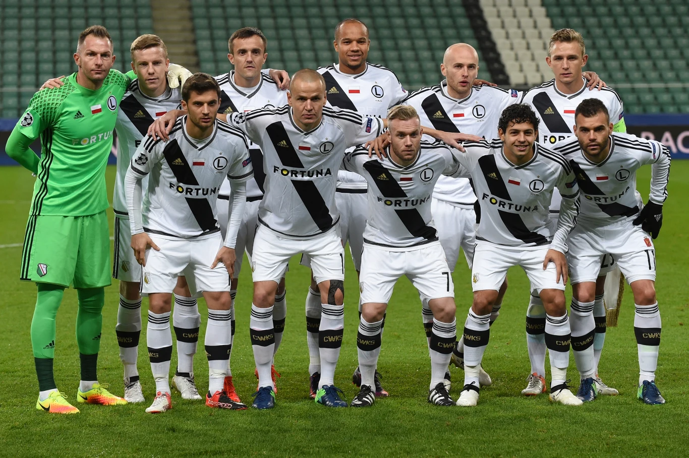 jedenastu piłkarzy drużyny Legia Warszawa w białych strojach z czarnym pasem i logiem sponsora, ustawionych w dwóch rzędach na murawie stadionu tuż przed rozpoczęciem meczu