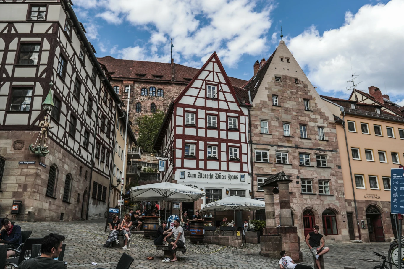 Rynek z zabytkowymi, szachulcowymi i kamiennymi kamienicami, centralnie usytuowana restauracja z białymi parasolami, ludzie siedzący przy stolikach oraz przechodnie na brukowanej ulicy, w tle fragmenty zamku i niebieskie niebo z chmurami.