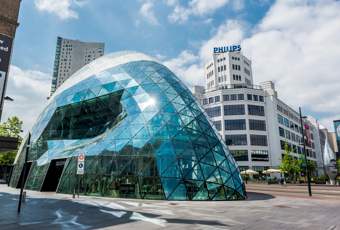 Nowoczesny budynek o futurystycznej, szklanej konstrukcji z licznymi trójkątnymi panelami dominuje na pierwszym planie, w tle widoczny jest biały gmach z napisem PHILIPS oraz wieżowiec, całość otoczona miejskim placem z niewielką ilością drzew i elemen...