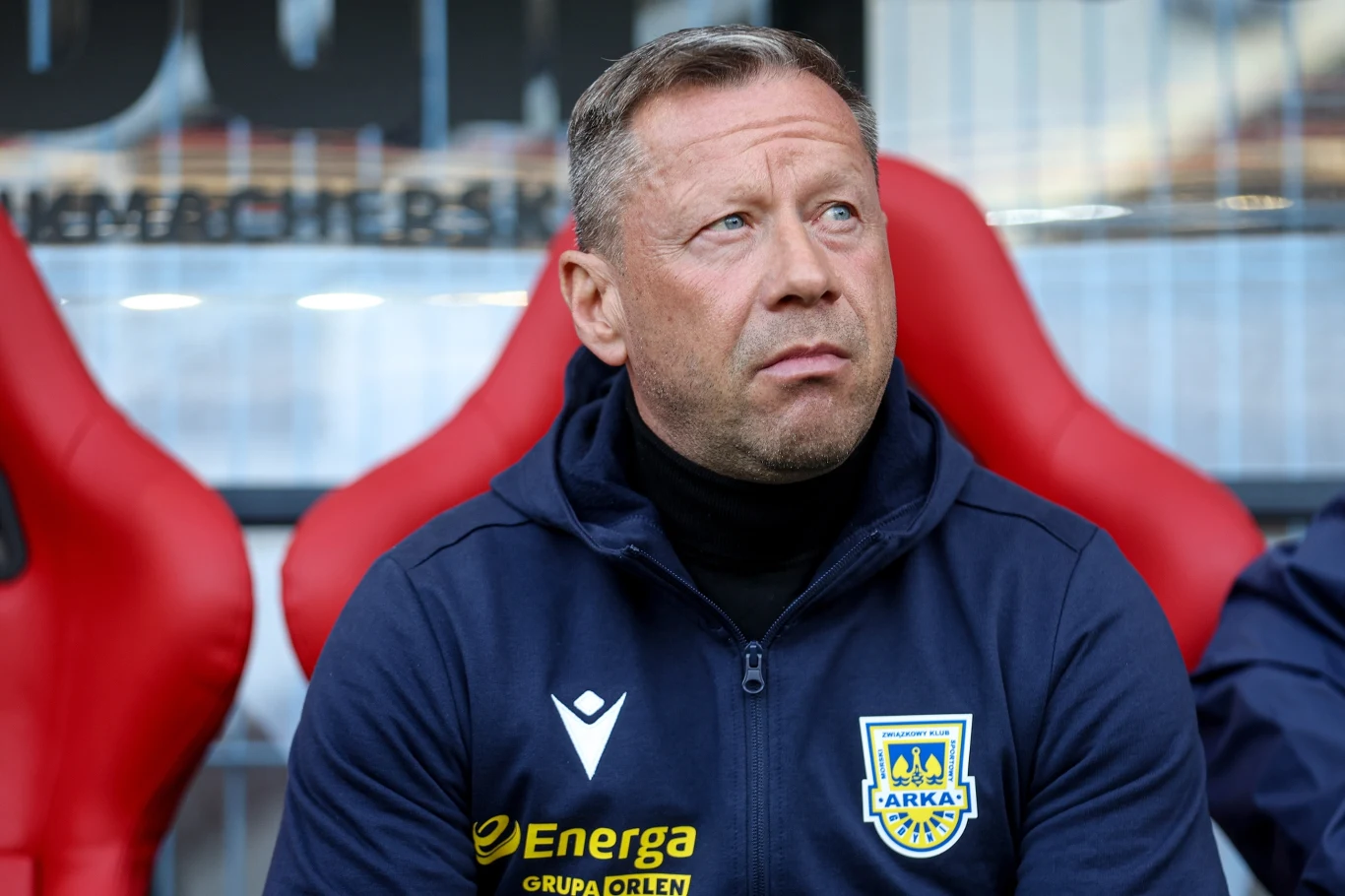 mężczyzna w kurtce z herbem Arki Gdynia oraz logotypami sponsorów siedzi na czerwonym fotelu stadionowym, wyrażając skupienie
