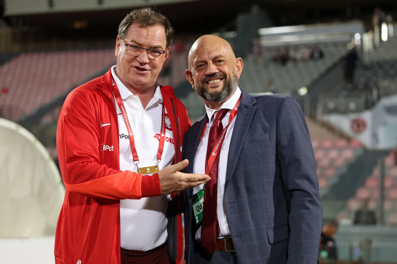 Dwóch mężczyzn stojących obok siebie na stadionie, jeden w sportowej kurtce reprezentacji Polski, drugi w garniturze z identyfikatorem na szyi, obaj uśmiechnięci i w serdecznym geście rozmowy.