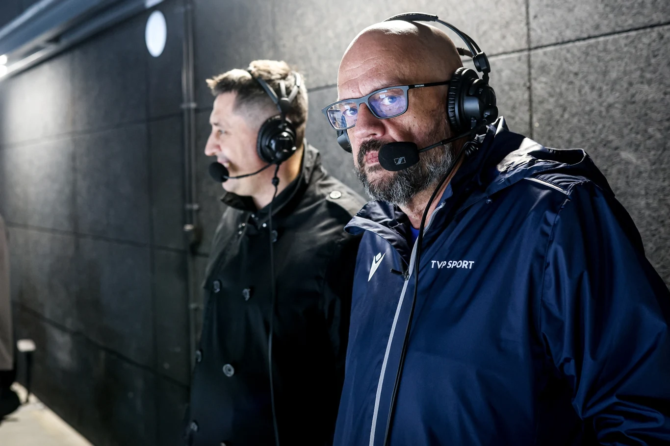 Dwóch komentatorów sportowych w słuchawkach z mikrofonami, jeden w kurtce TVP Sport, stojących w pomieszczeniu z ciemnymi ścianami.