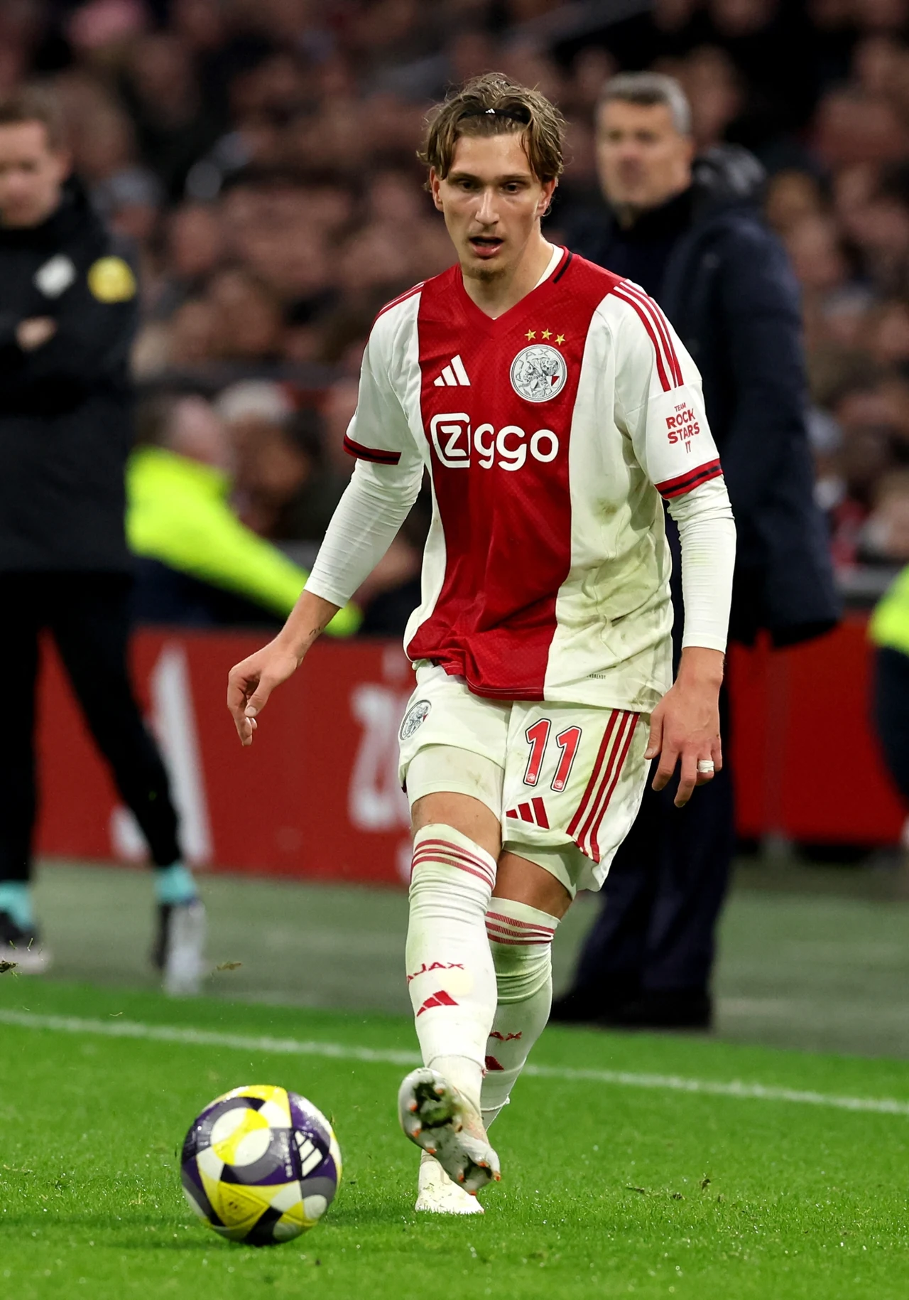 Piłkarz Ajaxu Amsterdam w biało-czerwonym stroju z numerem 11 prowadzi piłkę po murawie podczas meczu, wokół widoczni są sędziowie oraz kibice na trybunach.