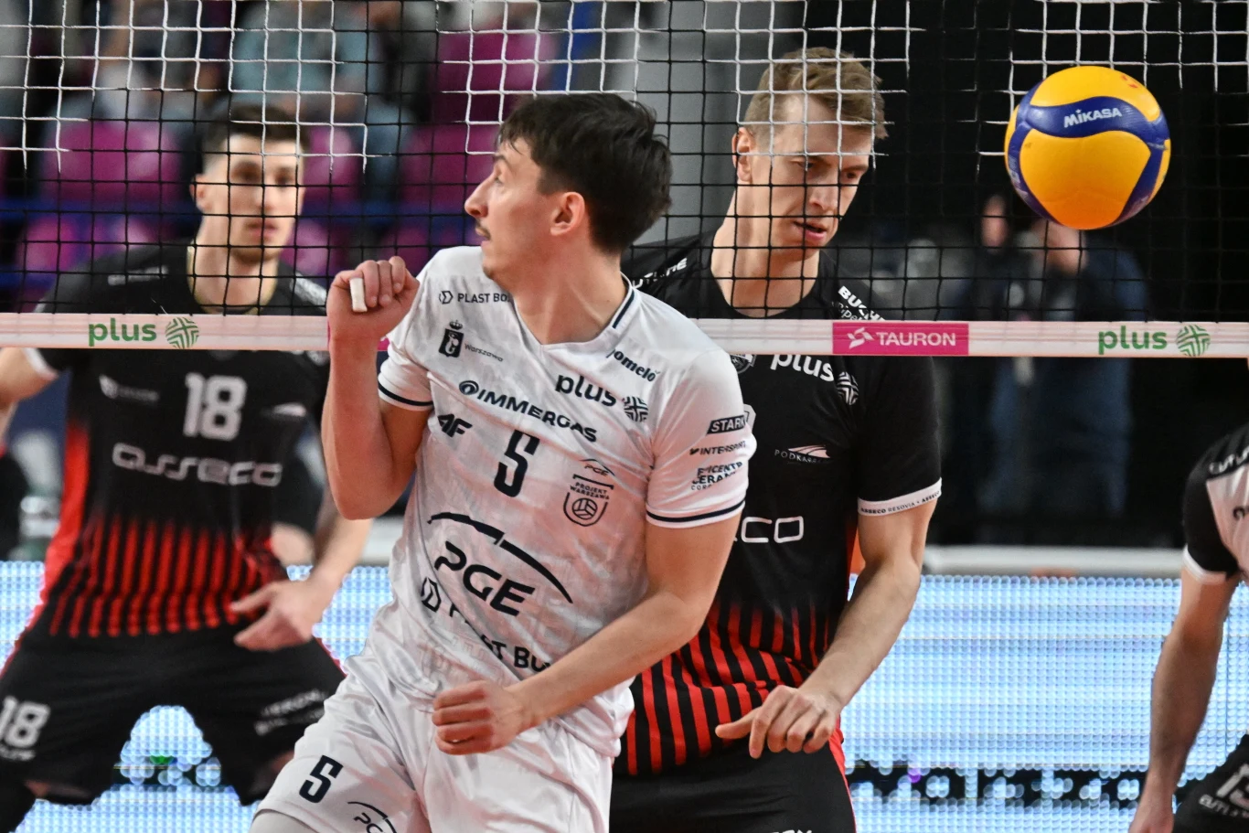 dwóch siatkarzy w trakcie gry przy siatce, jeden w białym stroju zwraca głowę w stronę lecącej piłki, drugi w czarnym stroju z rękami gotowymi do bloku, w tle rozmazani zawodnicy i siatka z widocznymi bandami reklamowymi