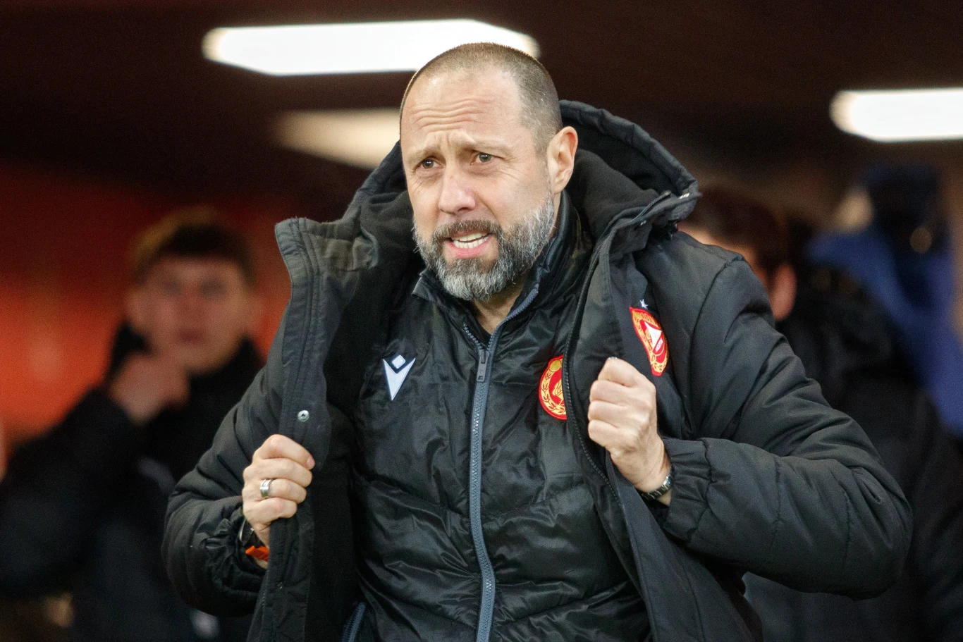 Igor Jovicević Igor Jovicević w czarnej kurtce sportowej z herbem klubu, trzyma kołnierz, patrzy przed siebie; w tle inne osoby.