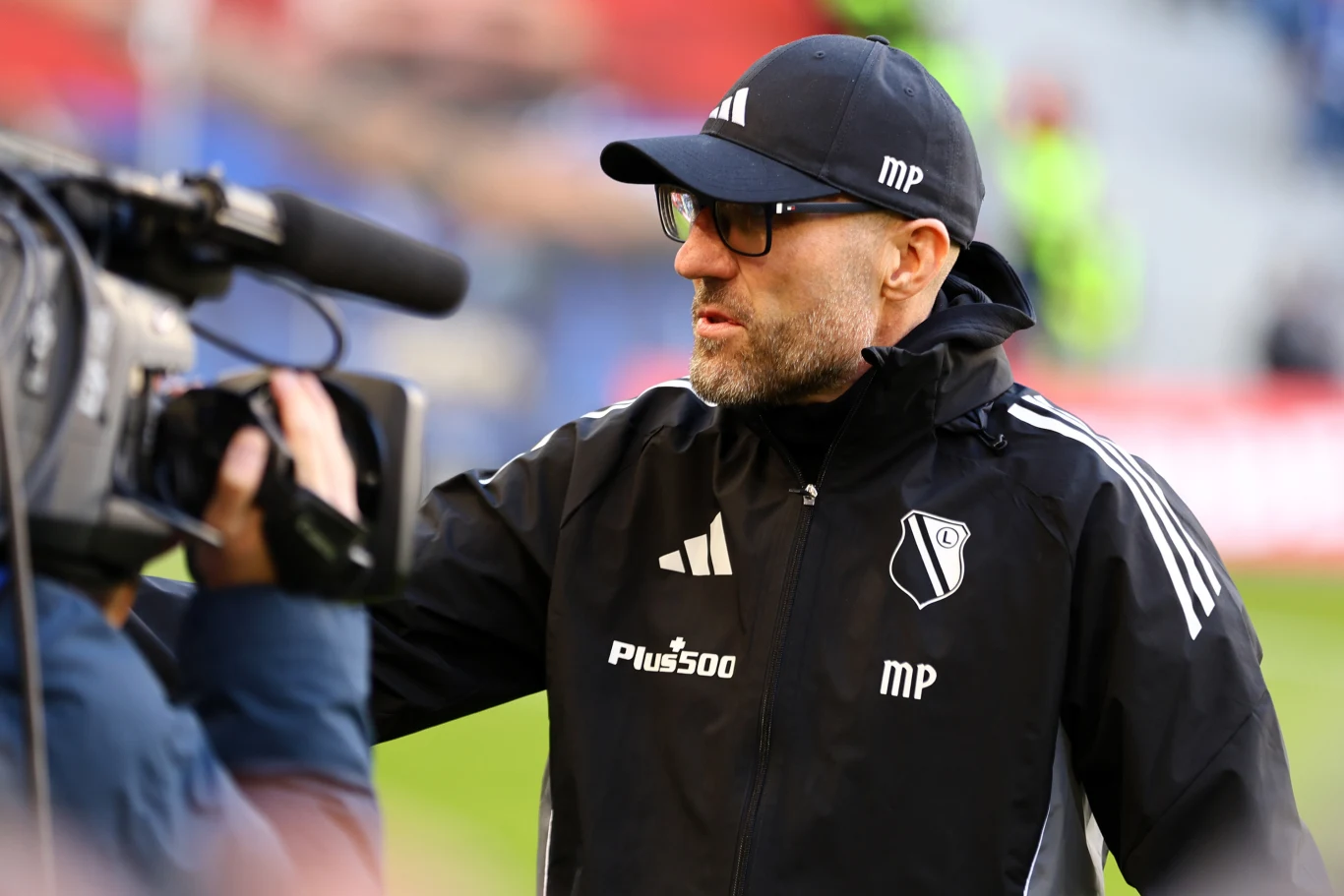 trener drużyny piłkarskiej Legia Warszawa w czarnej sportowej kurtce z logo klubu oraz czapce z daszkiem podczas udzielania wywiadu przed kamerą telewizyjną na stadionie