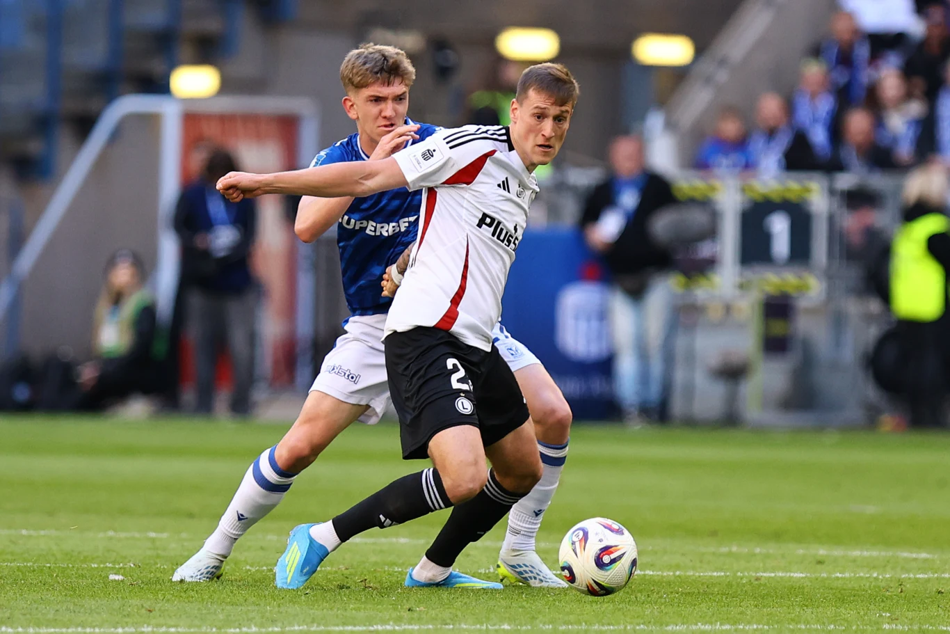 Legia starała się nękać Lecha wysokim pressingiem dwóch piłkarzy walczy o piłkę podczas meczu, jeden w niebieskim stroju próbuje odebrać piłkę rywalowi w białej koszulce, na drugim planie widać fragment boiska oraz rozmytych kibiców na trybunach