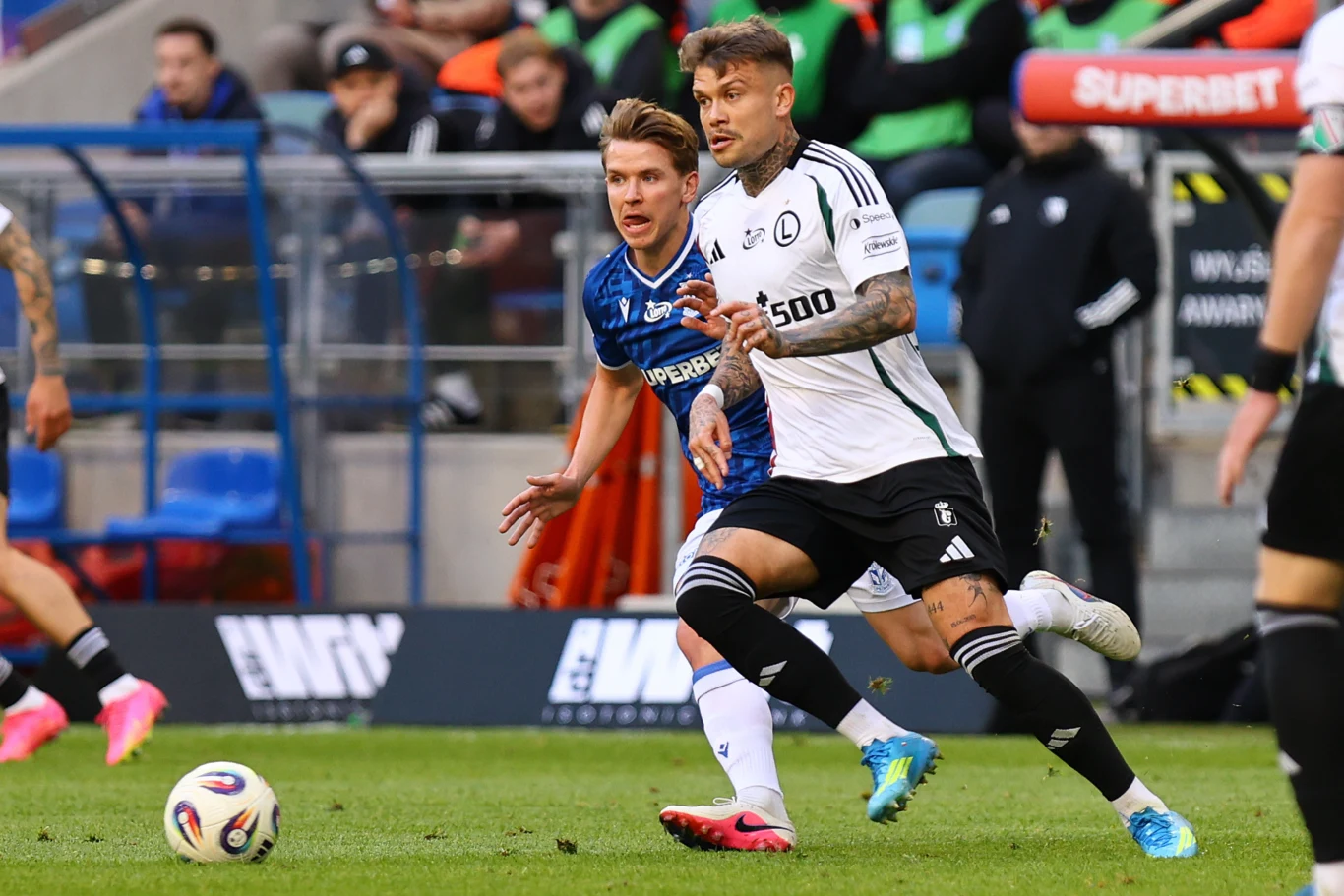 Lech Poznań - Legia Warszawa dwóch piłkarzy rywalizuje o piłkę podczas meczu, jeden w niebieskim stroju drużyny gospodarzy, drugi w biało-czarnym stroju zespołu gości, dynamiczna akcja na murawie stadionu, widoczni kibice i ławka rezerwowych w tle