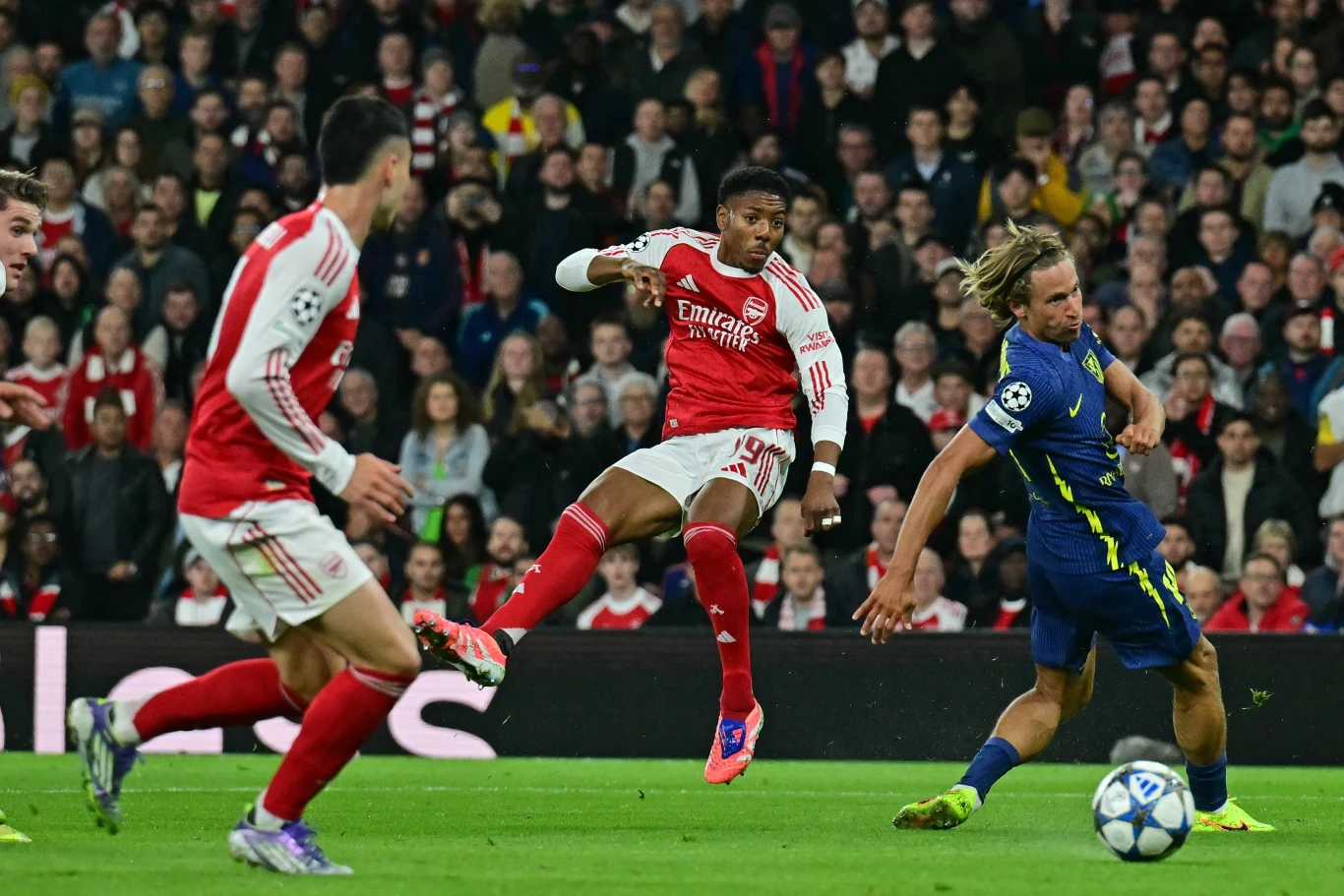 Mecz fazy ligowej Ligi Mistrzów Arsenal - Atletico Madryt w sezonie 2025/2026 Piłkarz w czerwono-białym stroju wykonuje dynamiczny strzał w kierunku bramki podczas meczu, podczas gdy zawodnik drużyny przeciwnej próbuje zablokować piłkę, a tłum kibiców w tle obserwuje akcję.