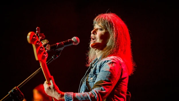 kobieta grająca na gitarze basowej i śpiewająca do mikrofonu podczas koncertu, oświetlona czerwonym światłem sceny