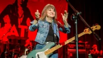 kobieta w dżinsowej kurtce stojąca na scenie z gitarą basową, w tle perkusja i sprzęt muzyczny, scena oświetlona czerwonym światłem