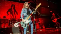 Wokalistka grająca na gitarze basowej stoi na środku sceny, ubrana w jasnoniebieski dżinsowy komplet, za nią perkusja, a w tle czerwony baner z czarną sylwetką postaci oraz nazwą zespołu, po prawej gitarzysta w trakcie występu.