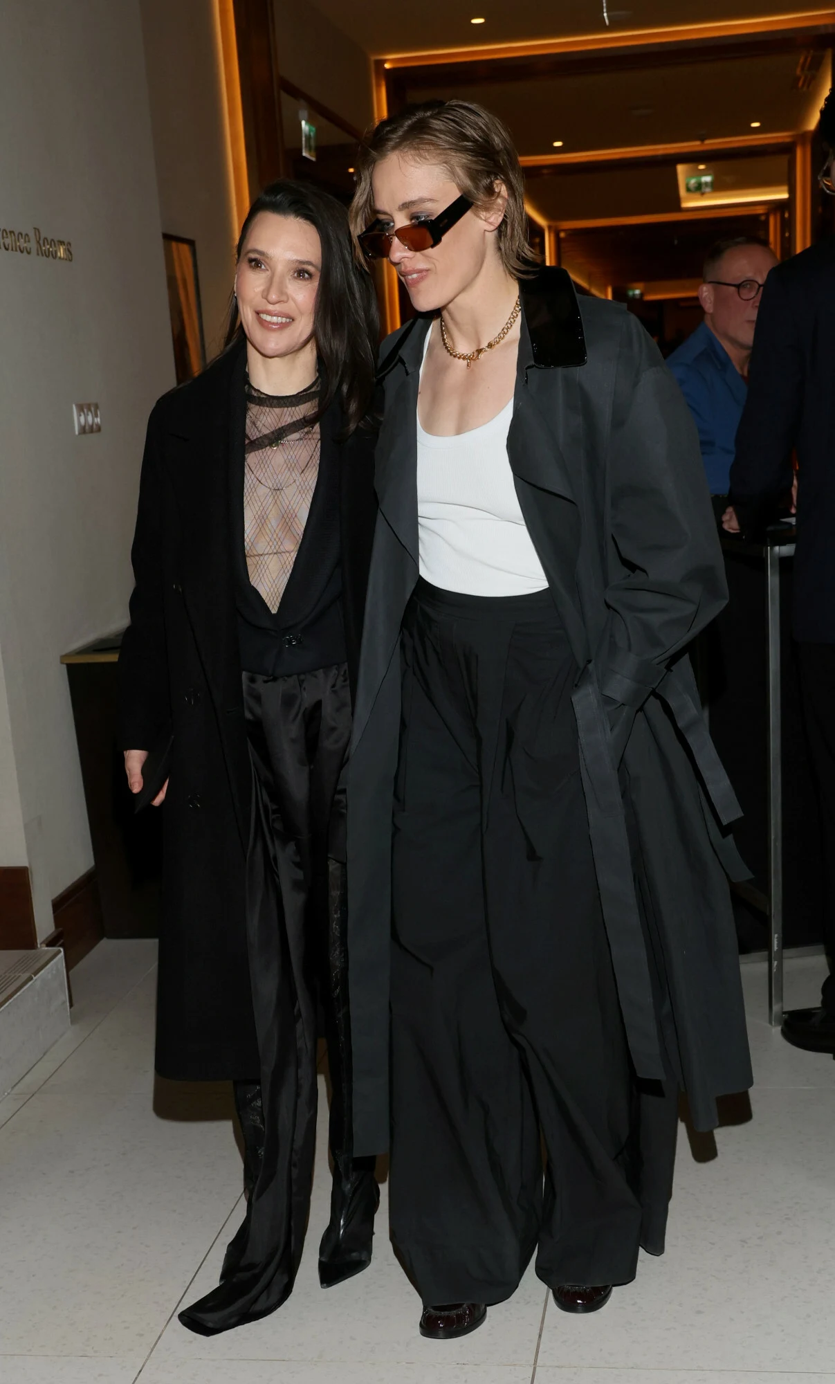 Dwie kobiety w eleganckich, ciemnych stylizacjach idą razem korytarzem; jedna ubrana w czarny garnitur z siateczkową bluzką, druga w szerokich spodniach, białym topie, długim płaszczu i okularach przeciwsłonecznych.