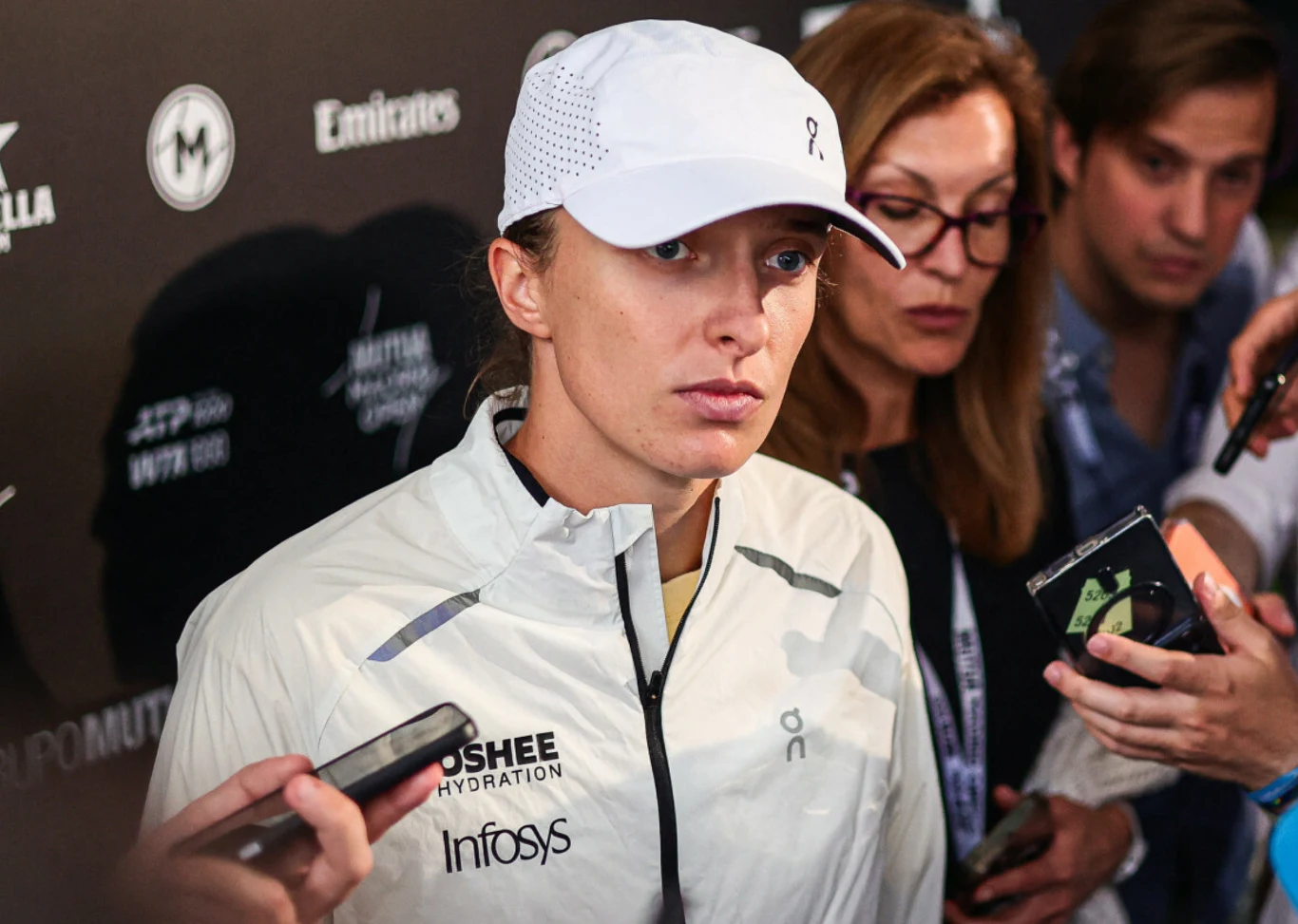 Iga Świątek sportowczyni ubrana w jasną kurtkę i białą czapkę z daszkiem odpowiada na pytania dziennikarzy, wokół niej widoczne są mikrofony i osoby z notatnikami oraz aparatami, tło stanowi ściana z logo sponsorów