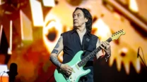 Mężczyzna z długimi ciemnymi włosami gra na jasnozielonej gitarze elektrycznej podczas koncertu; ma tatuaże na ramieniu i ubrany jest w ciemną, bezrękawną kamizelkę, na tle kolorowych, rozmazanych świateł scenicznych.