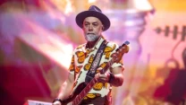 mężczyzna z brodą grający na gitarze basowej, ubrany w koszulę z motywem motyli oraz czarny kapelusz, na tle kolorowej, rozmytej scenografii z elementami instrumentów muzycznych