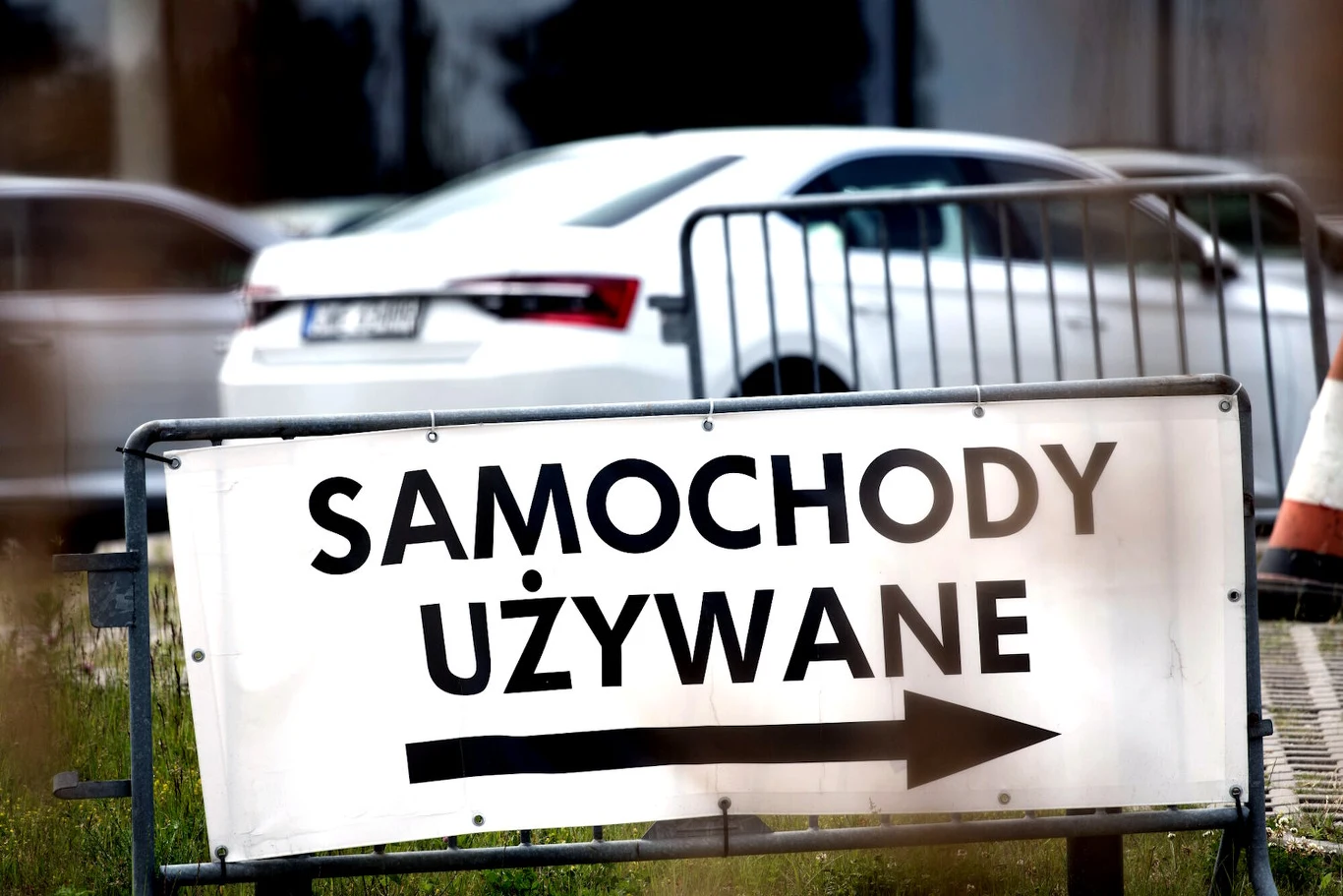 Systematycznie spada import używanych aut z zagranicy. Rynek przejmują dealerzy chińskich samochodów? Biała tablica z napisem 'SAMOCHODY UŻYWANE', strzałka w prawo, w tle białe auto osobowe przy ogrodzeniu.