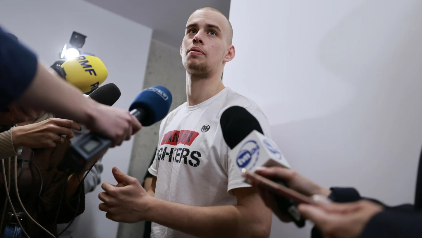 mężczyzna w białej koszulce z napisem FIGHTERS otoczony przez dziennikarzy z mikrofonami różnych stacji podczas wywiadu