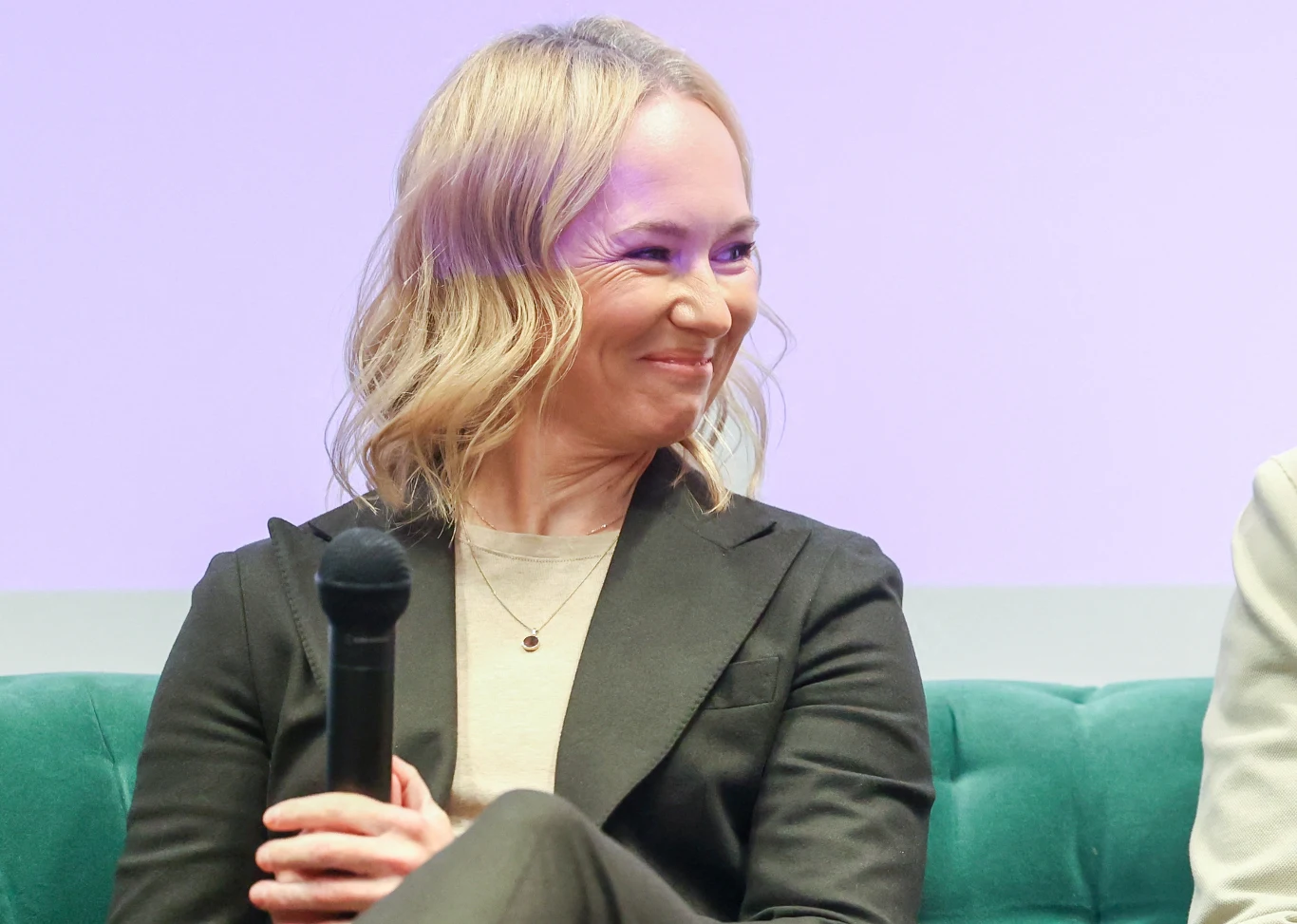 Kobieta w jasnych włosach ubrana w czarną marynarkę siedzi na zielonej sofie i trzyma mikrofon w dłoni, delikatnie się uśmiechając.