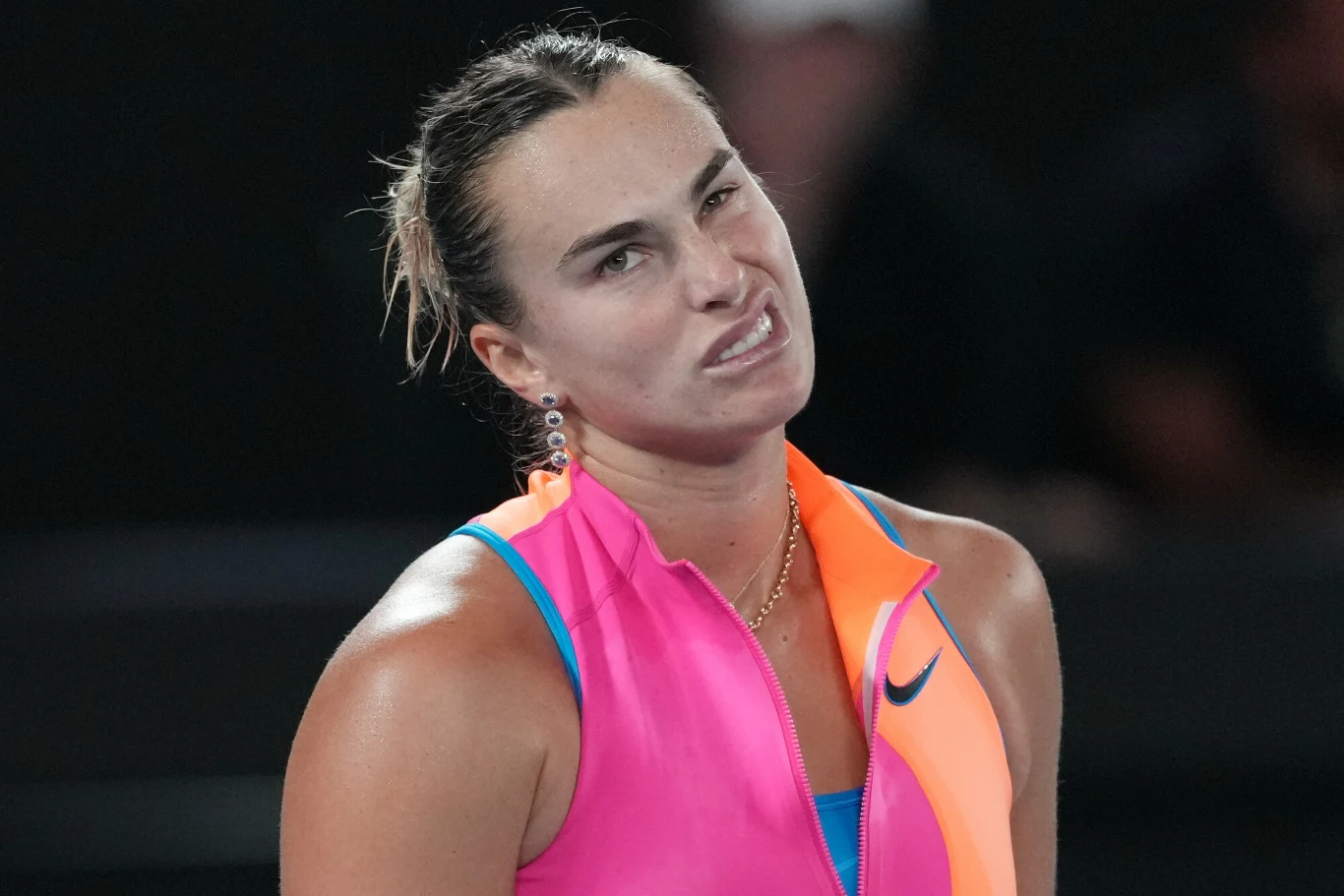 Kobieta w sportowym stroju w intensywnych kolorach różu i pomarańczu z rozpiętym kołnierzem, z wyraźnym grymasem niezadowolenia na twarzy oraz włosami związanymi w kucyk, stoi na tle ciemnego, rozmytego tła.