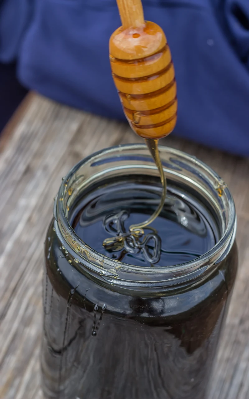 Szklany słoik wypełniony ciemnym miodem, nad którym unosi się drewniana łyżka do miodu z nabraną odrobiną gęstej, lejącej się substancji.