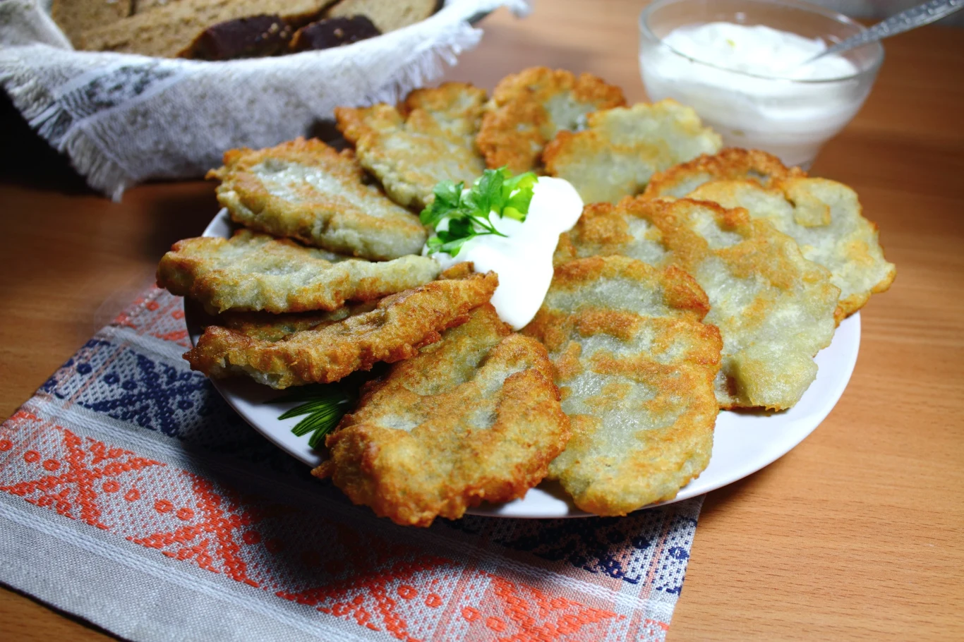 Placki ziemniaczane w wiejskiej wersji Wiejskie placki ziemniaczane na białym talerzu z natką pietruszki i śmietaną, w tle pieczywo i sos.