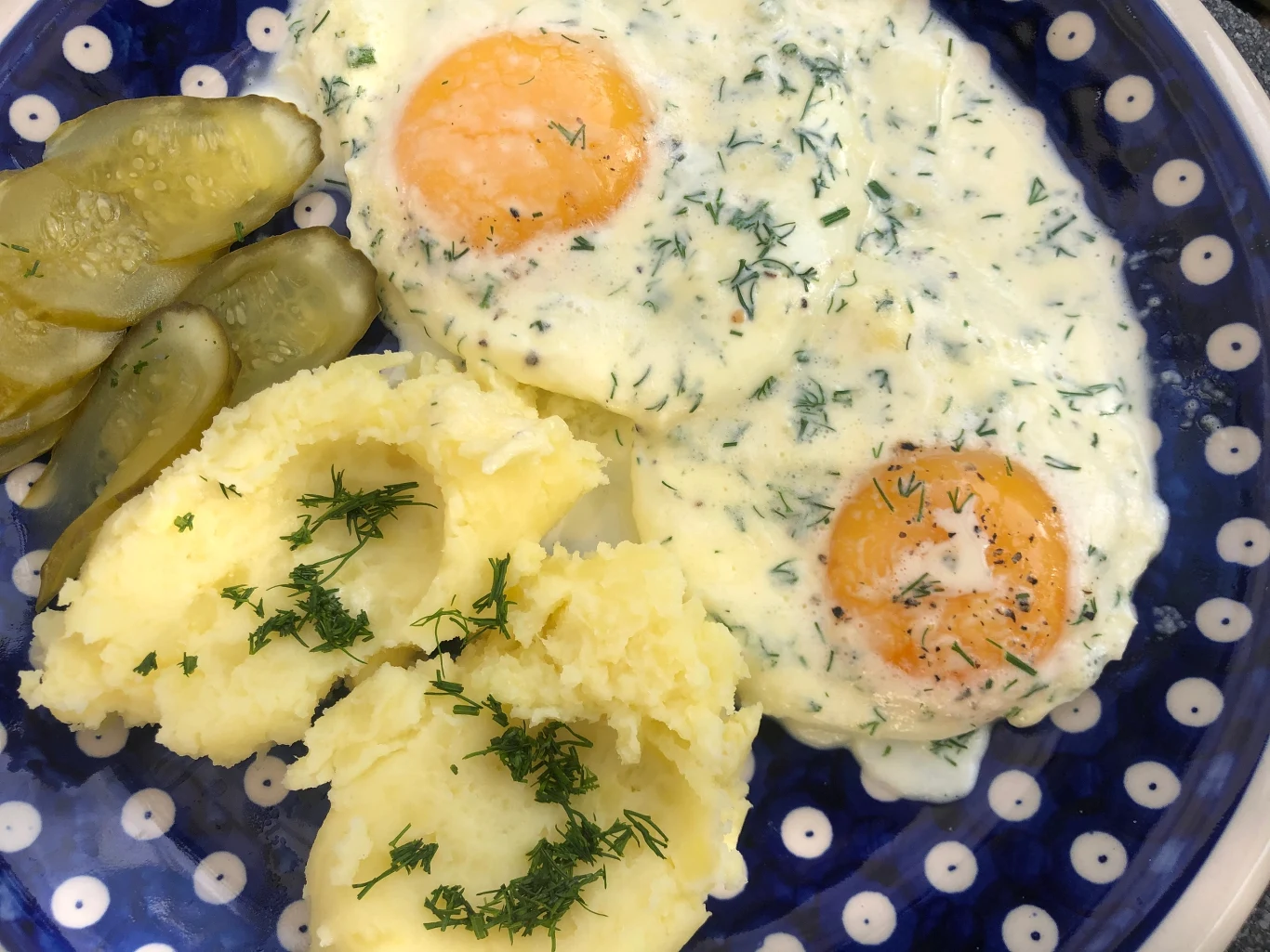 Jajka sadzone to pomysł na szybki obiad Dwa jajka w sosie koperkowym, porcje puree ziemniaczanego posypane świeżym koperkiem oraz plasterki ogórka kiszonego na granatowym talerzu w białe kropki.