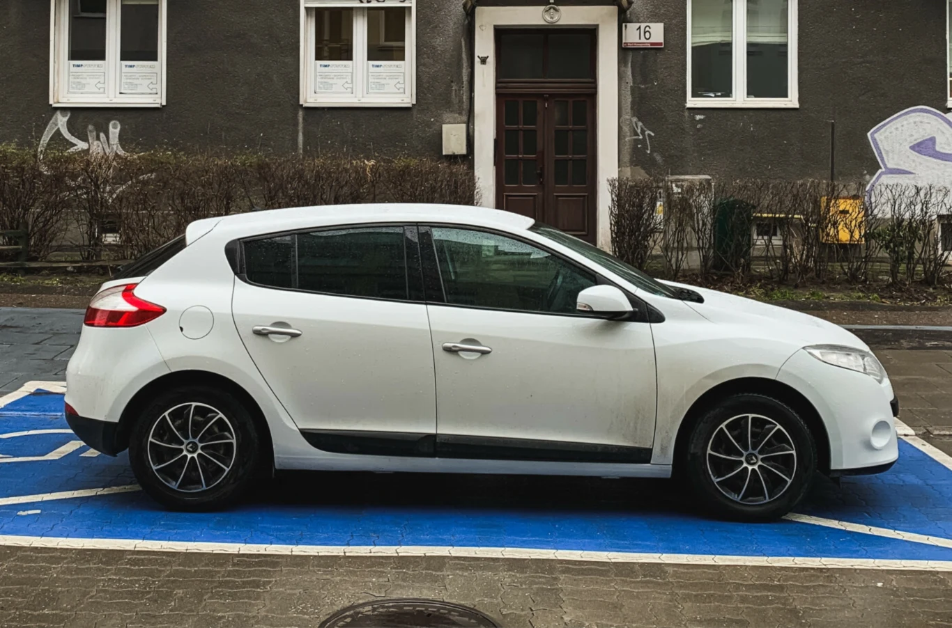 Podrabianie karty parkingowej - ile wynosi mandat i jaka kara grozi kierowcy? Biały samochód osobowy na miejscu dla osób z niepełnosprawnościami, w tle kamienica z drzwiami i oknami.