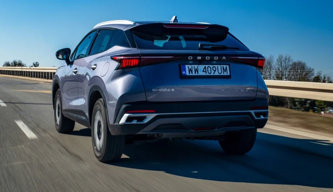 srebrny SUV Omoda C5 z polską rejestracją porusza się po pustej, dwupasmowej autostradzie w słoneczny dzień, na tle niebieskiego nieba i niskiej roślinności przy drodze