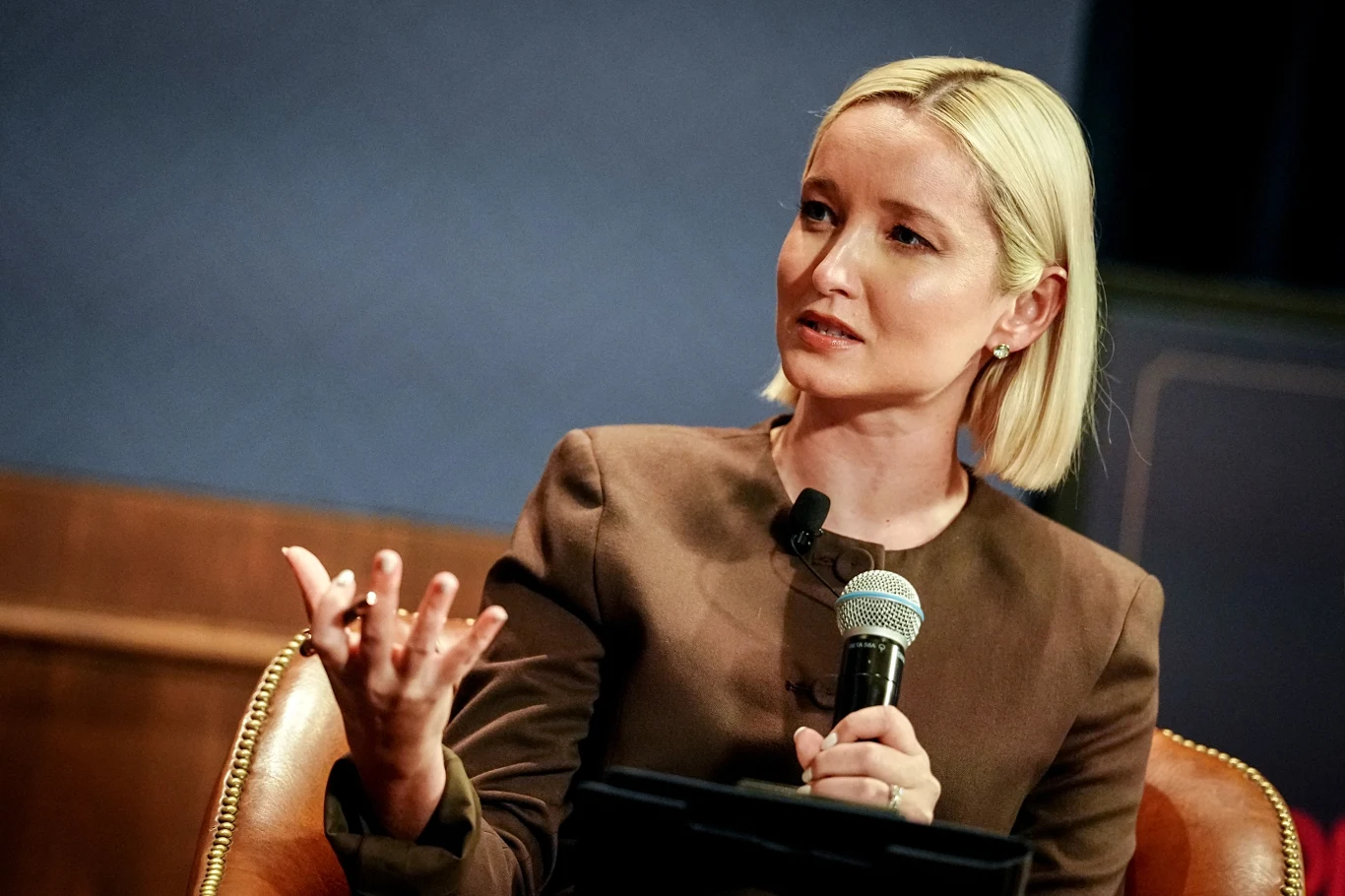 Dasha Burns, dziennikarka Politico Kobieta z blond włosami w eleganckiej, brązowej marynarce siedzi w skórzanym fotelu, trzyma mikrofon i gestykuluje dłonią podczas wypowiedzi, sugerując zaangażowanie w rozmowę.