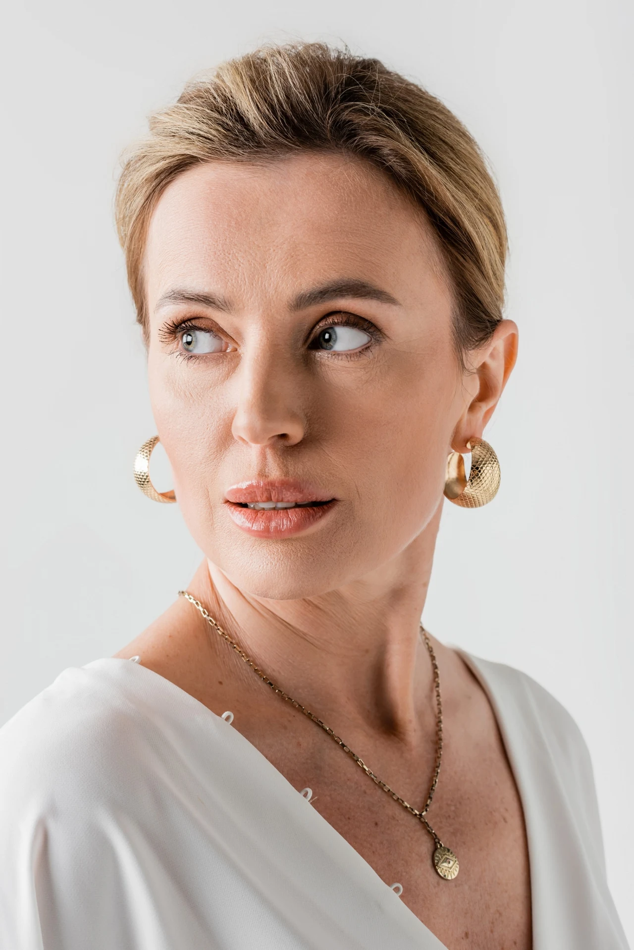 Kobieta o jasnych włosach upiętych do tyłu, ubrana w białą bluzkę, z dużymi złotymi kolczykami i delikatnym naszyjnikiem na szyi, spogląda w bok z poważnym wyrazem twarzy.