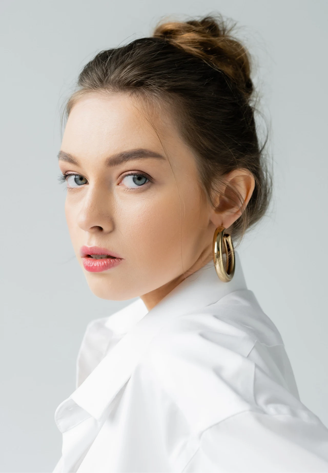 kobieta z upiętymi włosami, dużymi złotymi kolczykami kołami oraz subtelnym makijażem, ubrana w białą koszulę, patrzy w stronę obiektywu na jasnym tle