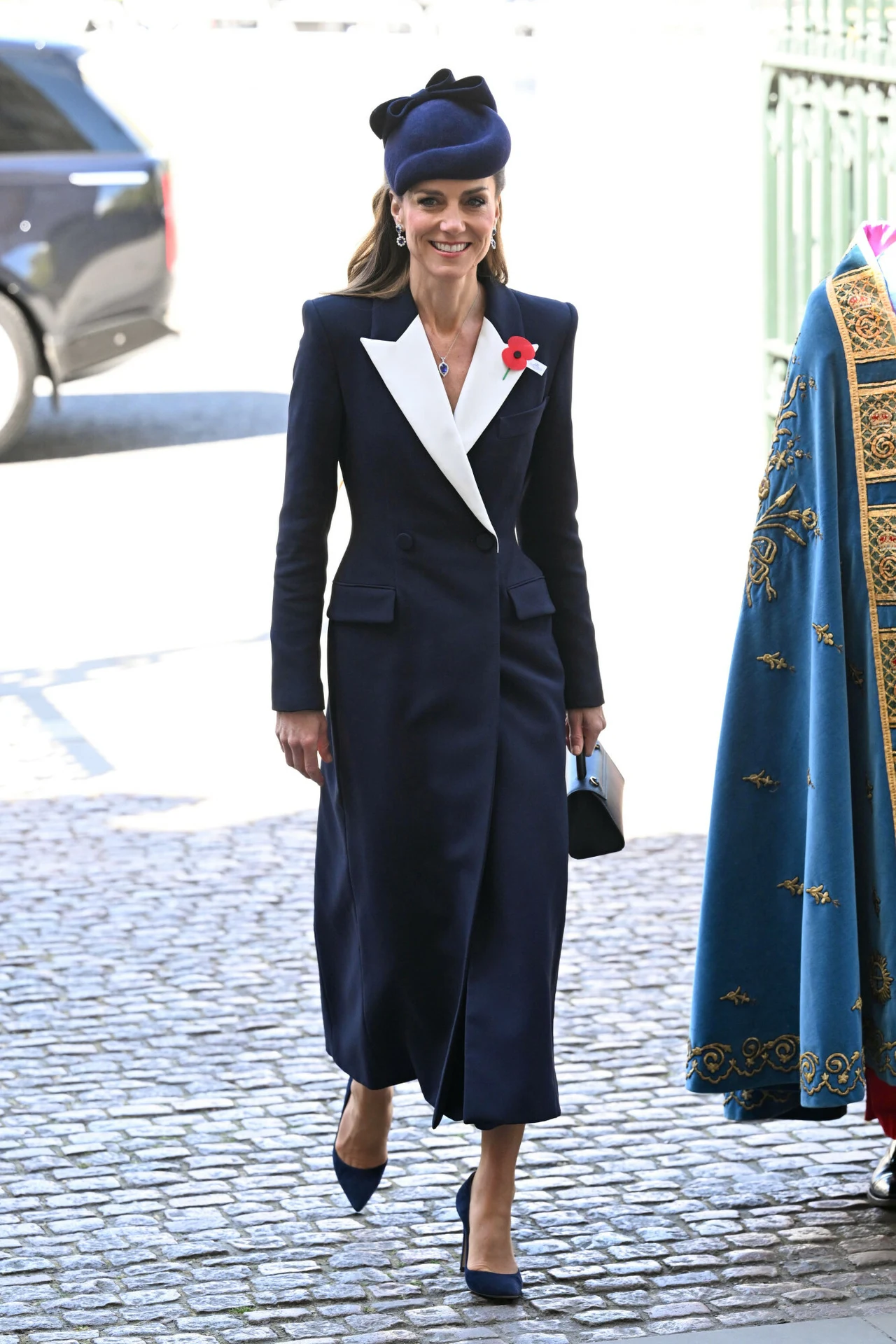 elegancka kobieta w granatowym płaszczu z białym kołnierzem, granatowych butach na obcasie i kapeluszu, trzymająca jasną torebkę i mająca przypięty czerwony kwiat na klapie