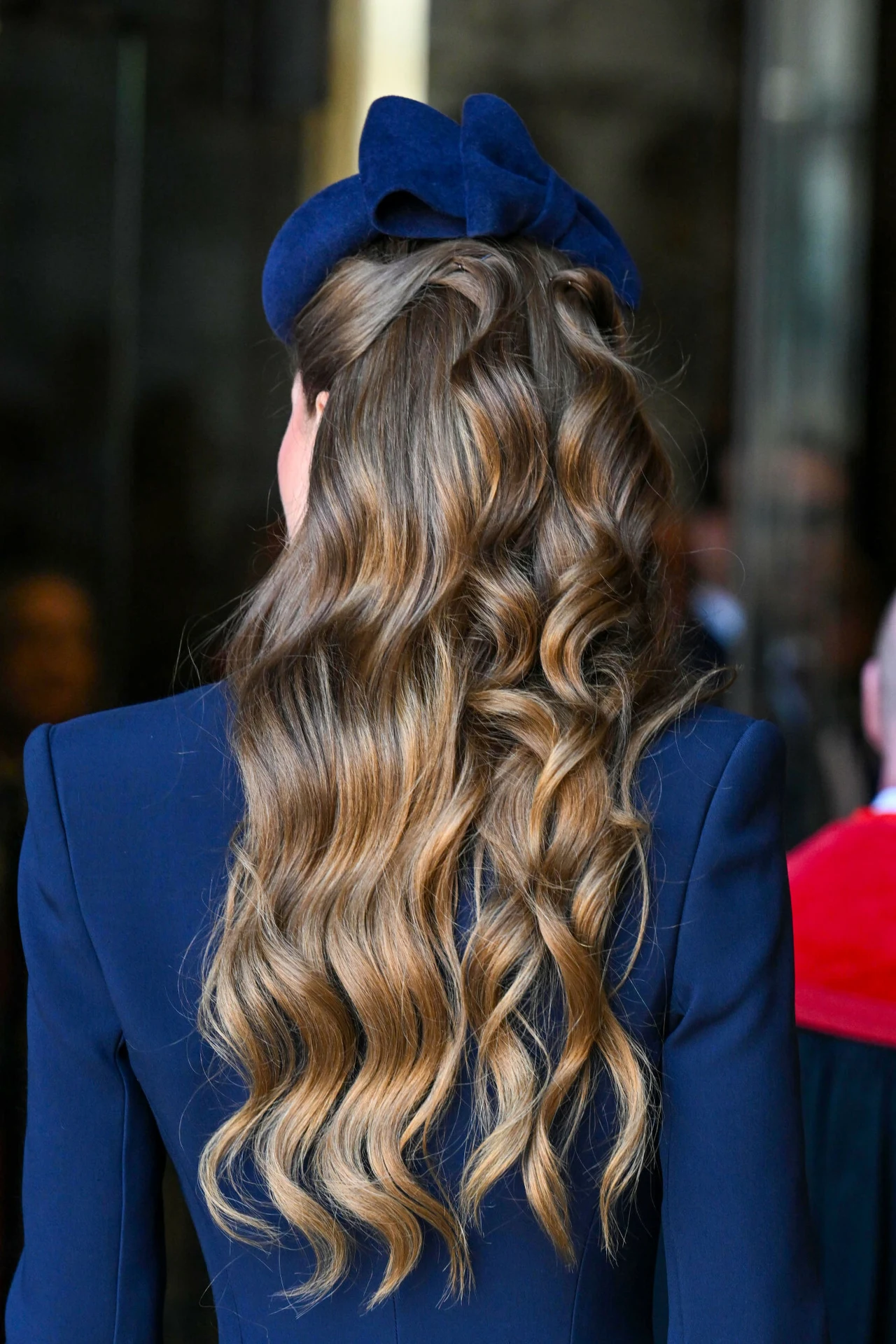 Długie, falowane brązowe włosy ozdobione granatową, aksamitną opaską w formie kokardy, elegancka granatowa marynarka podkreślająca formalny styl.