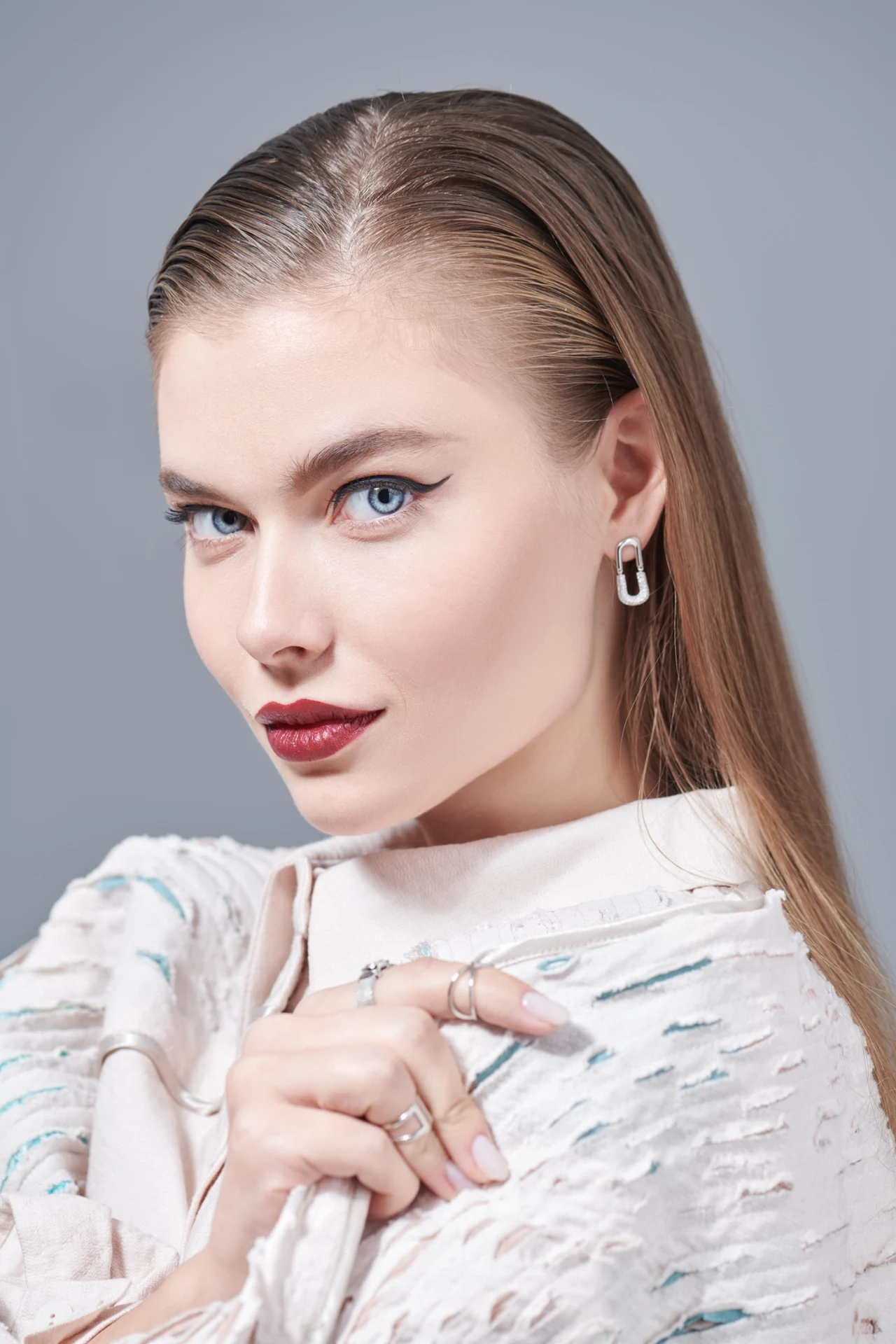 młoda kobieta o jasnej cerze i niebieskich oczach, z gładko zaczesanymi, prostymi włosami, ubrana w białą, teksturowaną bluzkę, nosząca srebrne kolczyki i kilka pierścionków, spoglądająca tajemniczo w stronę obiektywu na szarym tle