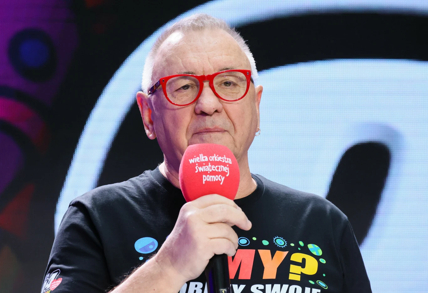 Jurek Owsiak ogłosił kolejne gwiazdy Pol'and'Rock Festivalu Jurek Owsiak w czerwonych okularach z mikrofonem WOŚP na tle jasnej, kolorowej grafiki podczas Pol'and'Rock Festivalu