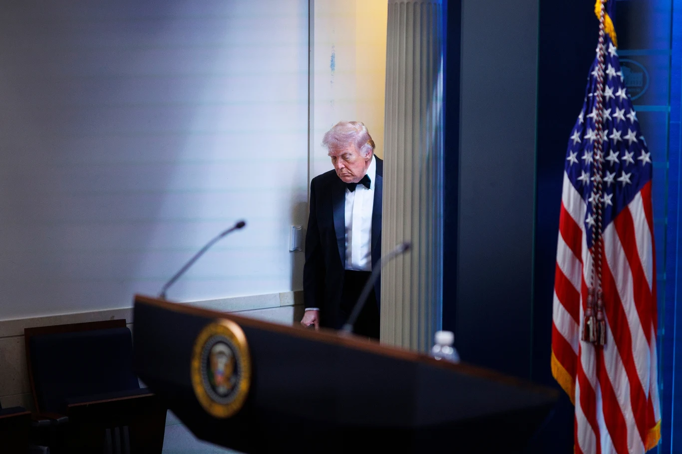 Donald Trump po strzelaninie podczas balu Donald Trump w smokingu wychodzi zza kotary; w tle pulpit z godłem USA i amerykańska flaga, Biały Dom