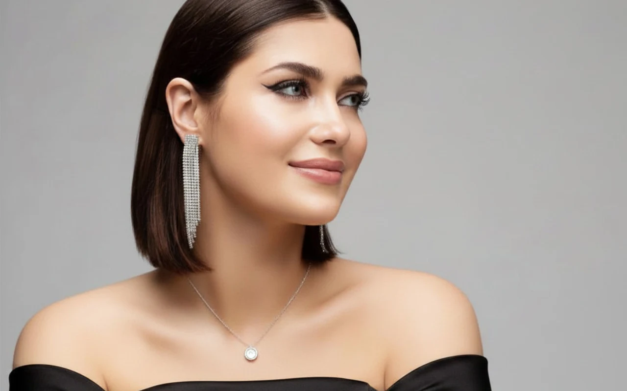 Młoda kobieta z krótkimi, ciemnymi włosami, lekko uśmiechnięta, ubrana w czarną, elegancką suknię z odkrytymi ramionami; podkreślona makijażem kreska na powiekach, błyszczące, długie kolczyki oraz delikatny naszyjnik z małą zawieszką.