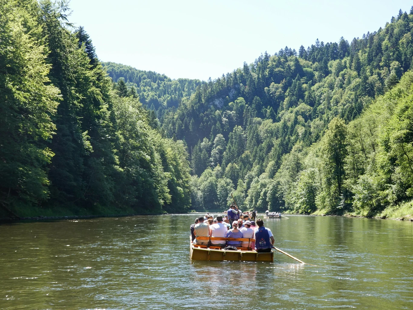 Spływ Dunajcem to pomysł na jednodniową wycieczkę w majówkę. Grupa osób na drewnianej tratwie podczas spływu Dunajcem, rzeka otoczona zielonymi lasami i wzgórzami.