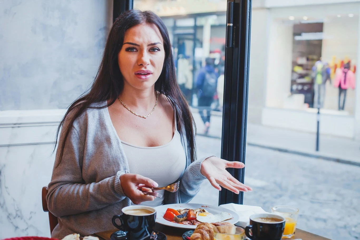 Zwykle coś im nie pasuje: jedzenie w restauracji, praca, widok z okna, kolejki w sklepie. Zmęczenie w takim towarzystwie gwarantowane kobieta o długich, ciemnych włosach siedzi przy stoliku w kawiarni, zdezorientowana lub zdenerwowana, z gestem dłoni sugerującym niezadowolenie; przed nią na stole filiżanka kawy, talerze z jedzeniem oraz sok pomarańczowy, w tle przez okno widać ulicę ...