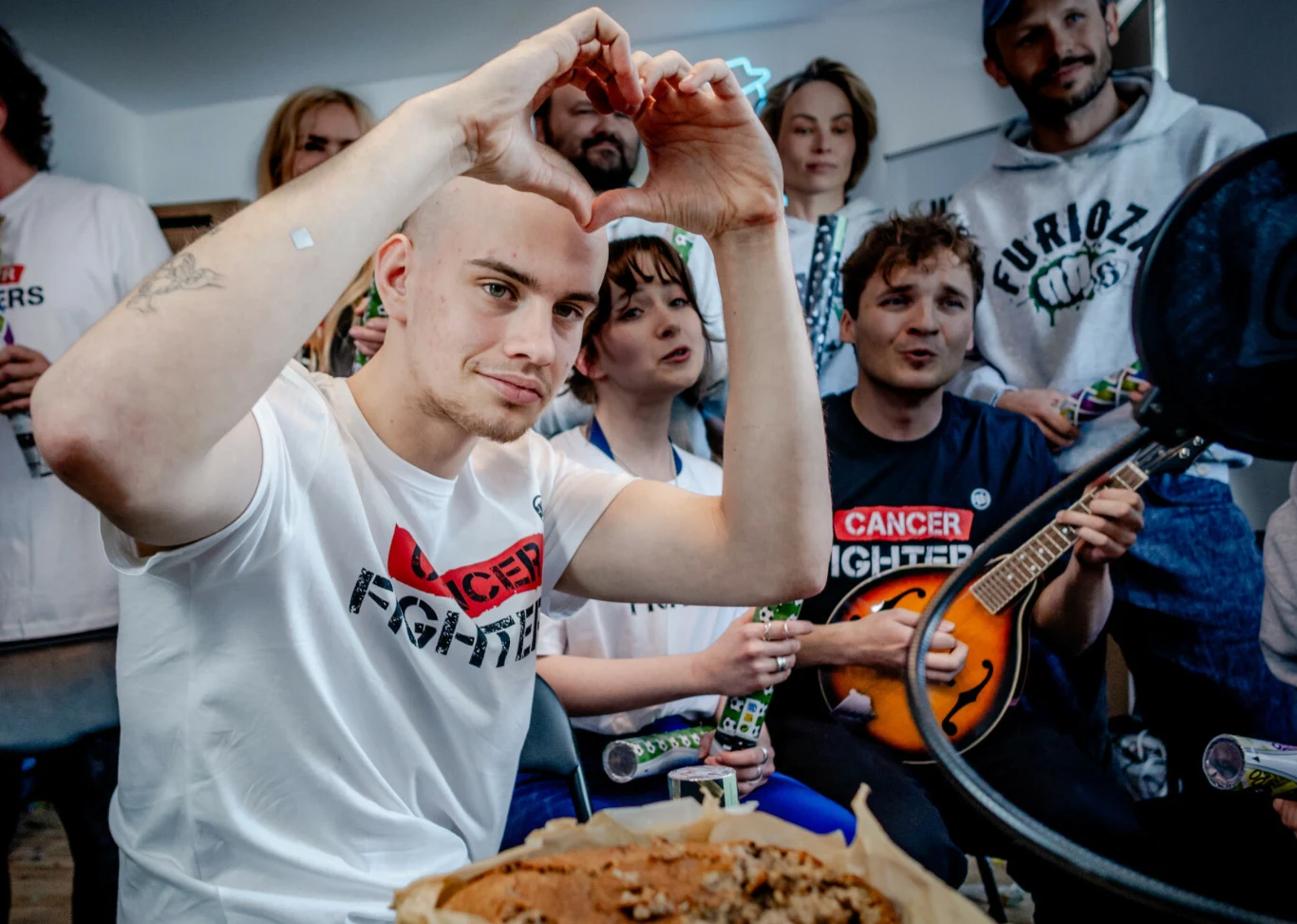grupa młodych osób z instrumentami muzycznymi i napojami, w centrum łysy mężczyzna w koszulce z napisem 'CANCER FIGHTERS' pokazuje gest serca dłońmi, w tle rozśpiewani przyjaciele, na pierwszym planie fragment ciasta