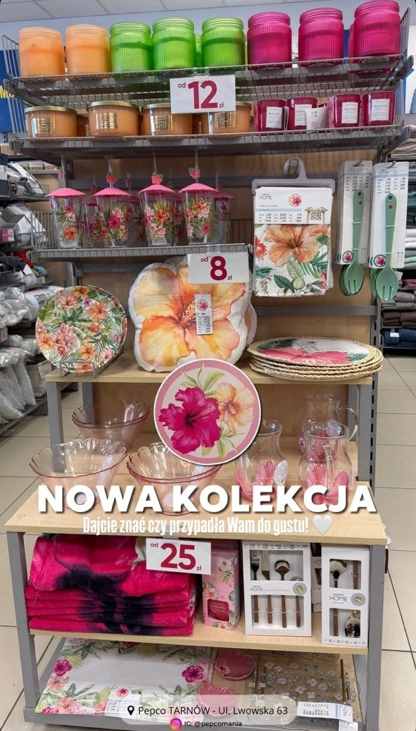 regał sklepowy z akcesoriami domowymi ozdobionymi motywami kwiatowymi i tropikalnymi, w tym poduszki, talerze, pojemniki, kubki oraz naczynia w różowo-zielonej kolorystyce, na środku duży napis 'NOWA KOLEKCJA'
